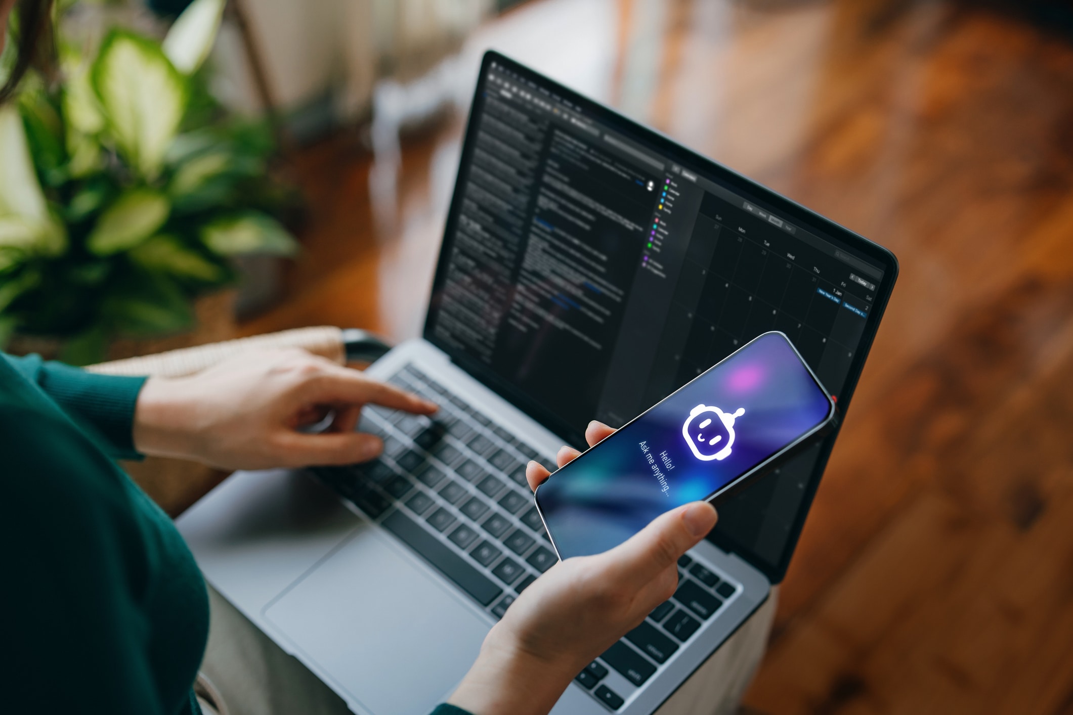 A person sits with a laptop on their lap, coding on the screen while holding a smartphone showing an AI chatbot icon and “Say Hello to AI”. A plant and wooden floor are visible in the background.