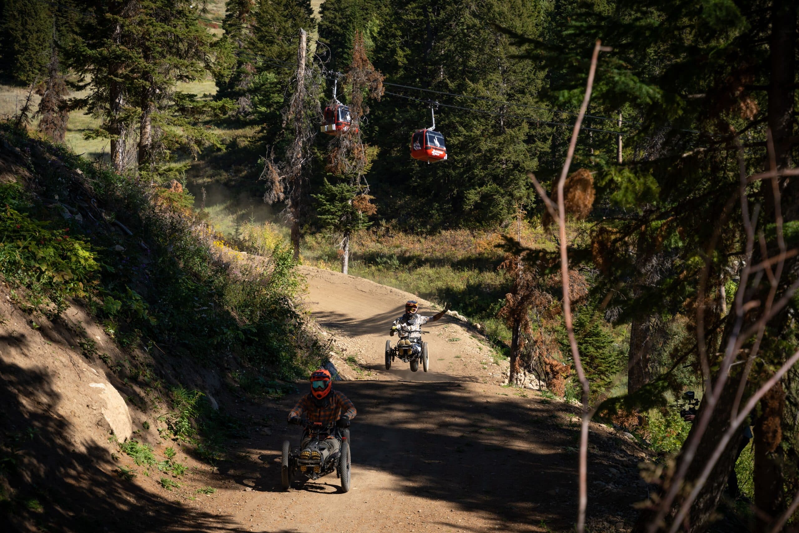 Three people ride adaptive mountain bikes on a dirt trail through a forested area, with red cable cars visible overhead and sunlight filtering through the trees.