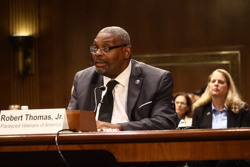 A man in a suit and glasses, identified by a nameplate as Robert Thomas, Jr., speaks at a government hearing. Seated people and a wood-paneled room are visible in the background.