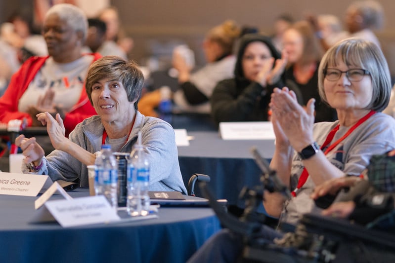 People sit at round tables, clapping and smiling during an event. In the foreground, two women with name tags are applauding, with water bottles and papers on the table in front of them. Other attendees are visible in the background.