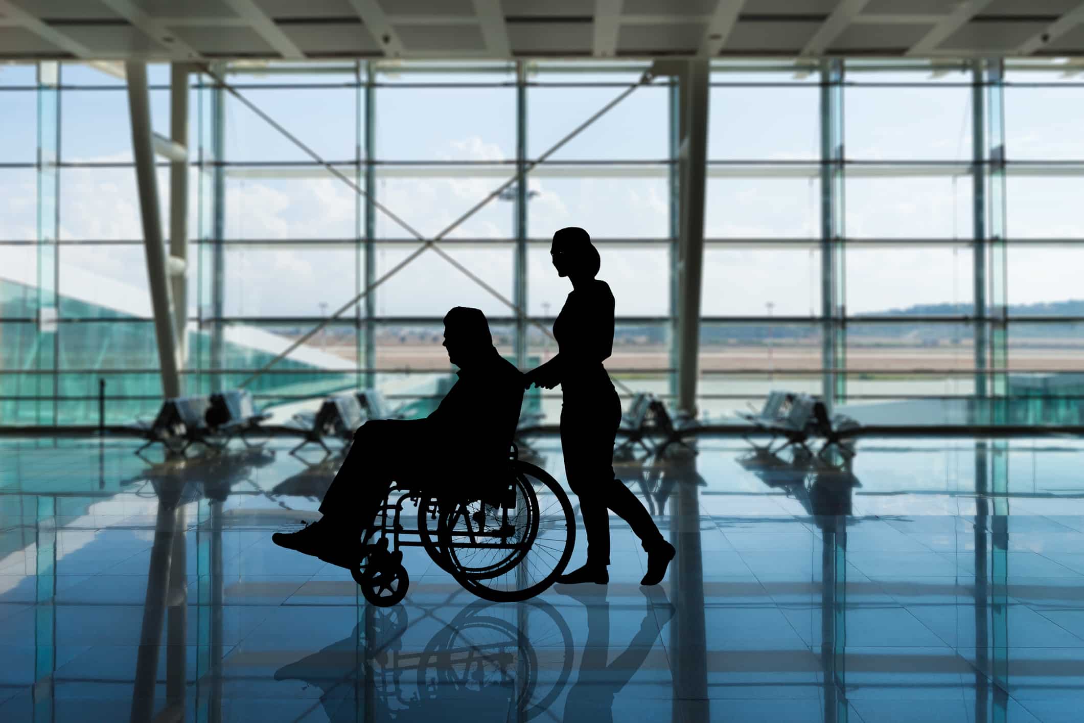 Silhouette of a person in a wheelchair being pushed by another person in an airport terminal, with large windows showing the runway and seats in the background.