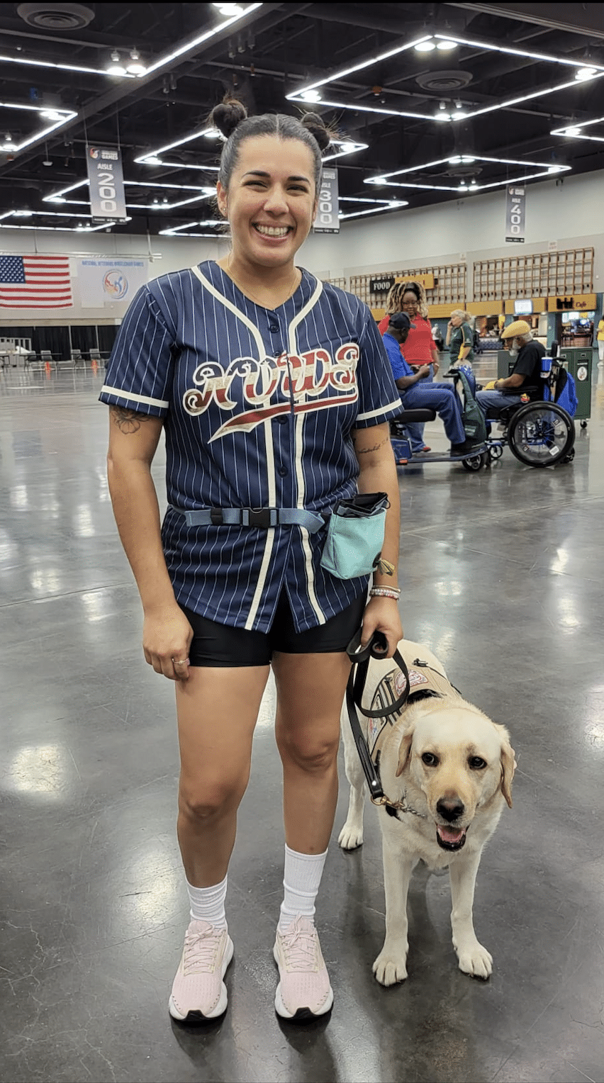 A smiling person in shorts, a striped "Rudes" jersey, and pink sneakers stands indoors next to a yellow Labrador service dog wearing a harness. People and wheelchairs are visible in the background.