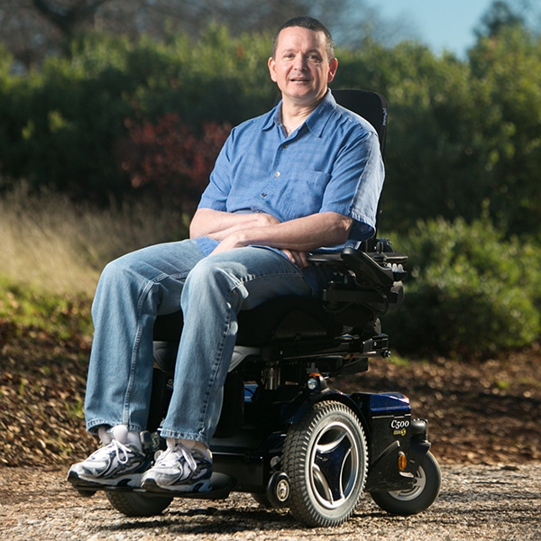A man wearing a blue shirt and jeans smiles while sitting in a motorized wheelchair outdoors, with greenery and trees in the background.