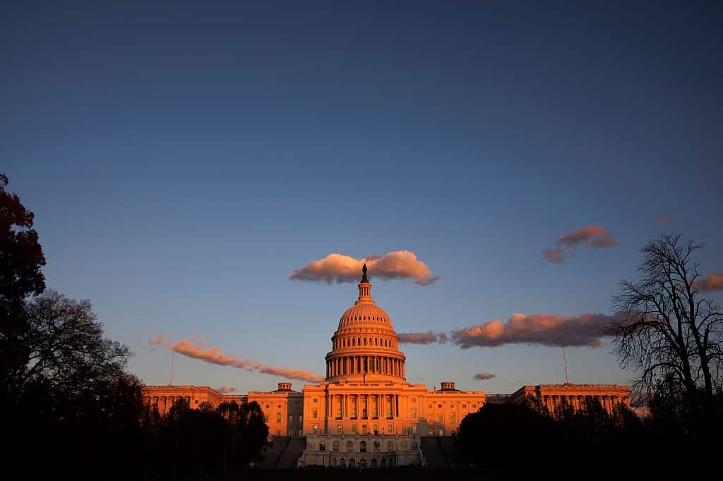 The U.S. Capitol building is illuminated by warm sunlight at sunset, with a clear sky and scattered clouds above, and trees silhouetted in the foreground.