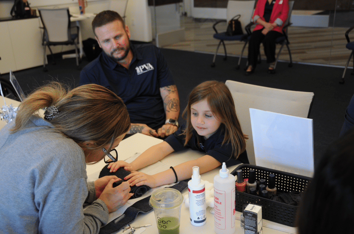 A young girl sits at a table getting her nails painted by a woman, while a man with tattoos watches beside her. Nail supplies and bottles are on the table. Other people are seated in the background.