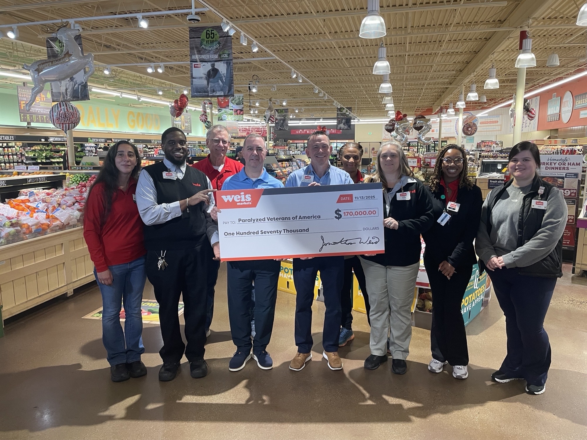 A group of nine people standing in a grocery store, holding an oversized check for $170,000 made out to "Four-Footed Veterans of America" from Weis Markets. Shelves of groceries are visible in the background.