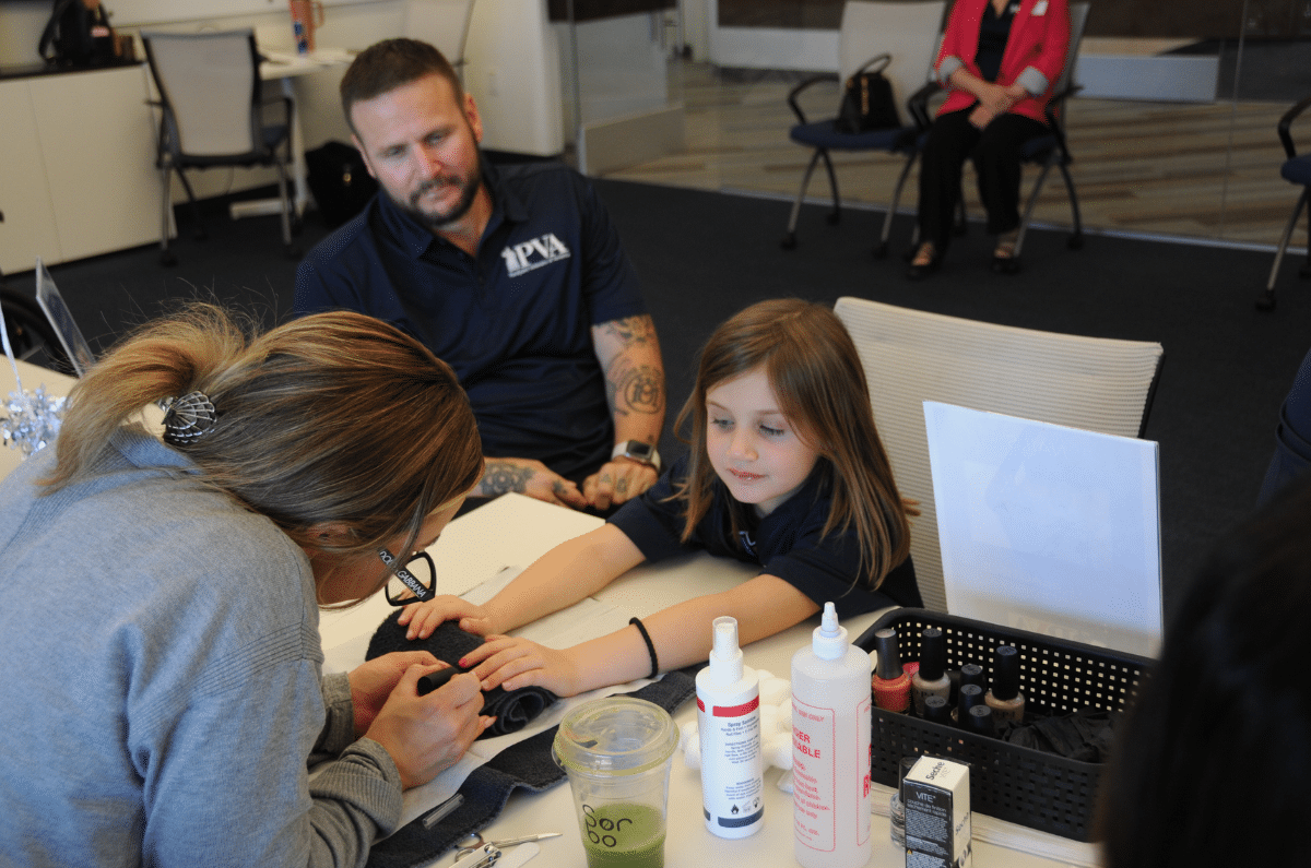A woman paints a young girl's nails at a table, while a man sits nearby watching. Bottles of nail supplies are on the table, and another woman is seated in the background. The scene appears casual and friendly.