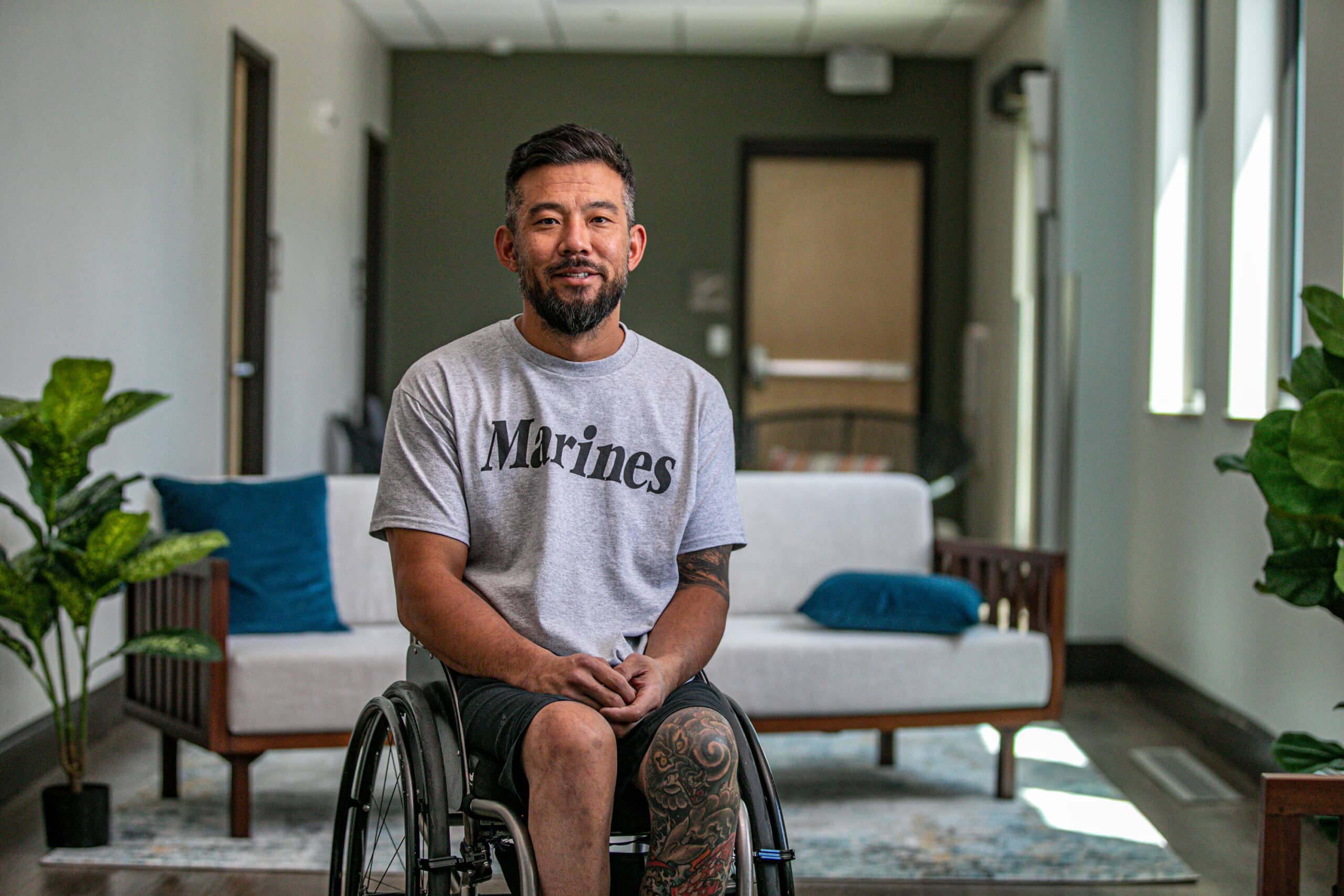 A man in a wheelchair wearing a gray "Marines" t-shirt sits smiling in a well-lit, modern room with plants, a white couch, and blue cushions in the background.