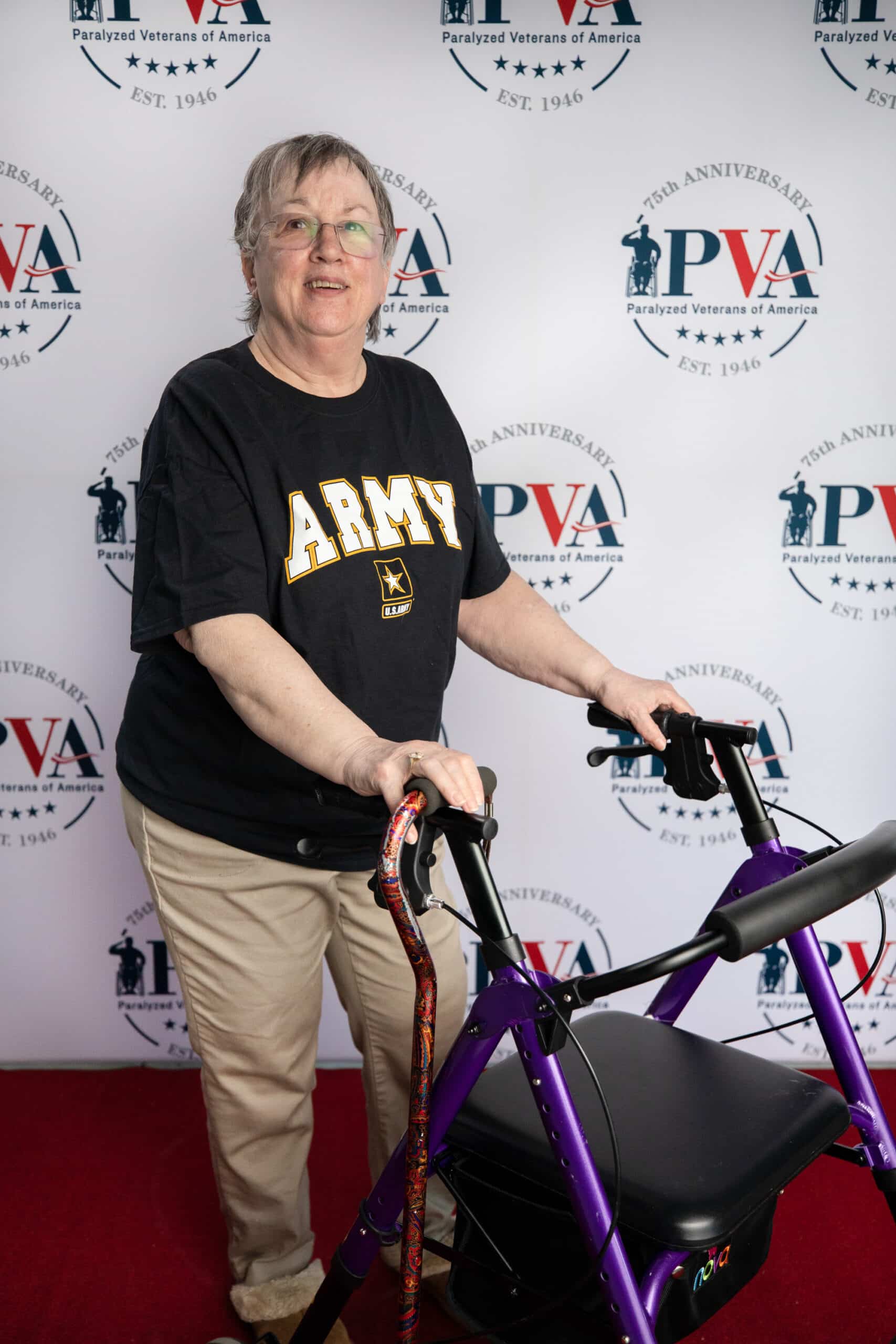 An older woman wearing an "Army" t-shirt smiles while using a purple walker, standing in front of a backdrop with "Paralyzed Veterans of America" logos and their 75th anniversary branding.