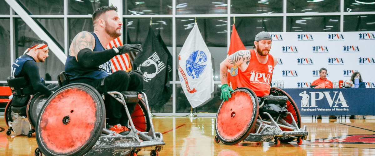 Three male athletes in wheelchairs compete in an indoor wheelchair rugby game. Two wear red jerseys, one wears blue, and sports flags and banners hang in the background. The court is shiny and reflects the action.