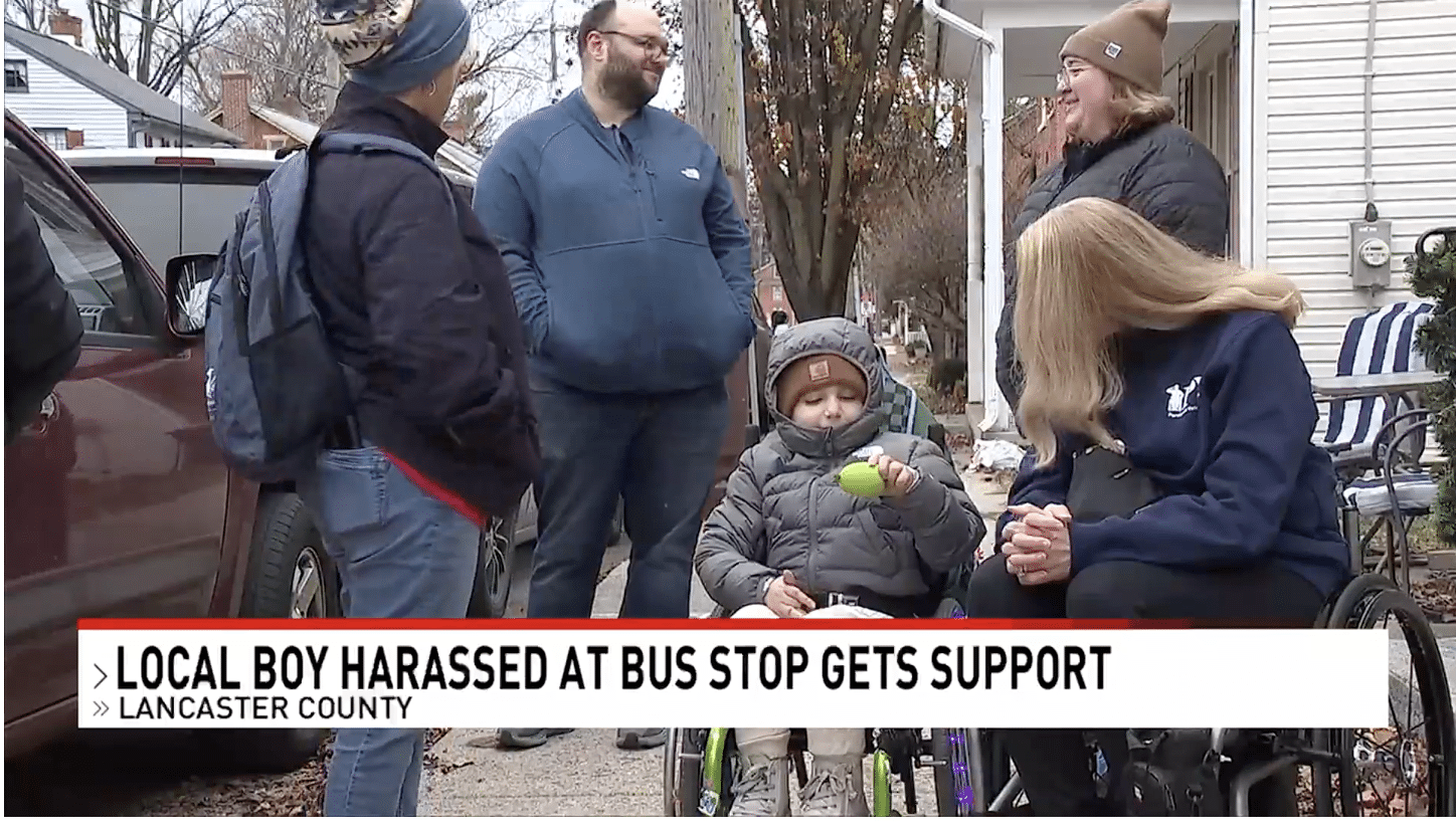 A group of people, including a young boy in a wheelchair, gather outdoors on a sidewalk. The text overlay reads, "Local boy harassed at bus stop gets support – Lancaster County.