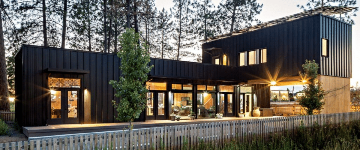 A modern black metal house with large windows sits among tall pine trees, illuminated warmly at dusk. A wooden fence and small trees line the front yard.