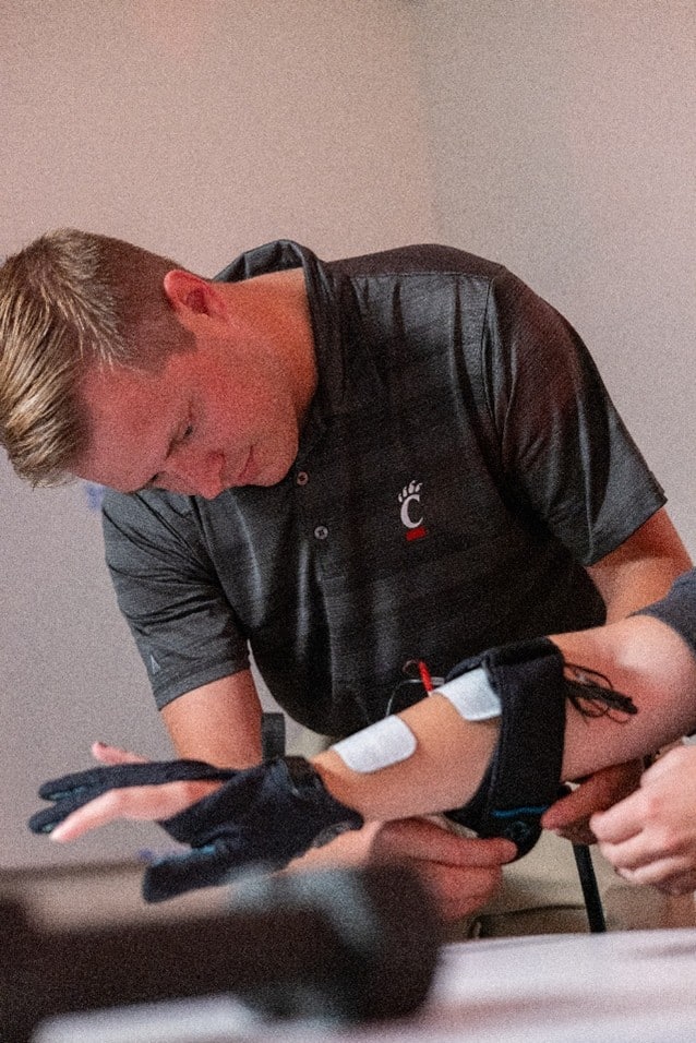 a man in a dark polo shirt is adjusting a wearable device with sensors on another person's forearm and hand, for a medical or research purpose, in an indoor setting.
