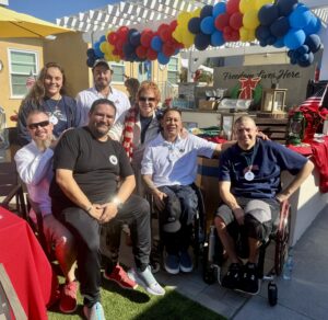 A group of seven people, including two in wheelchairs, smile together at an outdoor party with colorful balloons and a “Freedom Lives Here” sign in the background. The setting is festive and sunny.