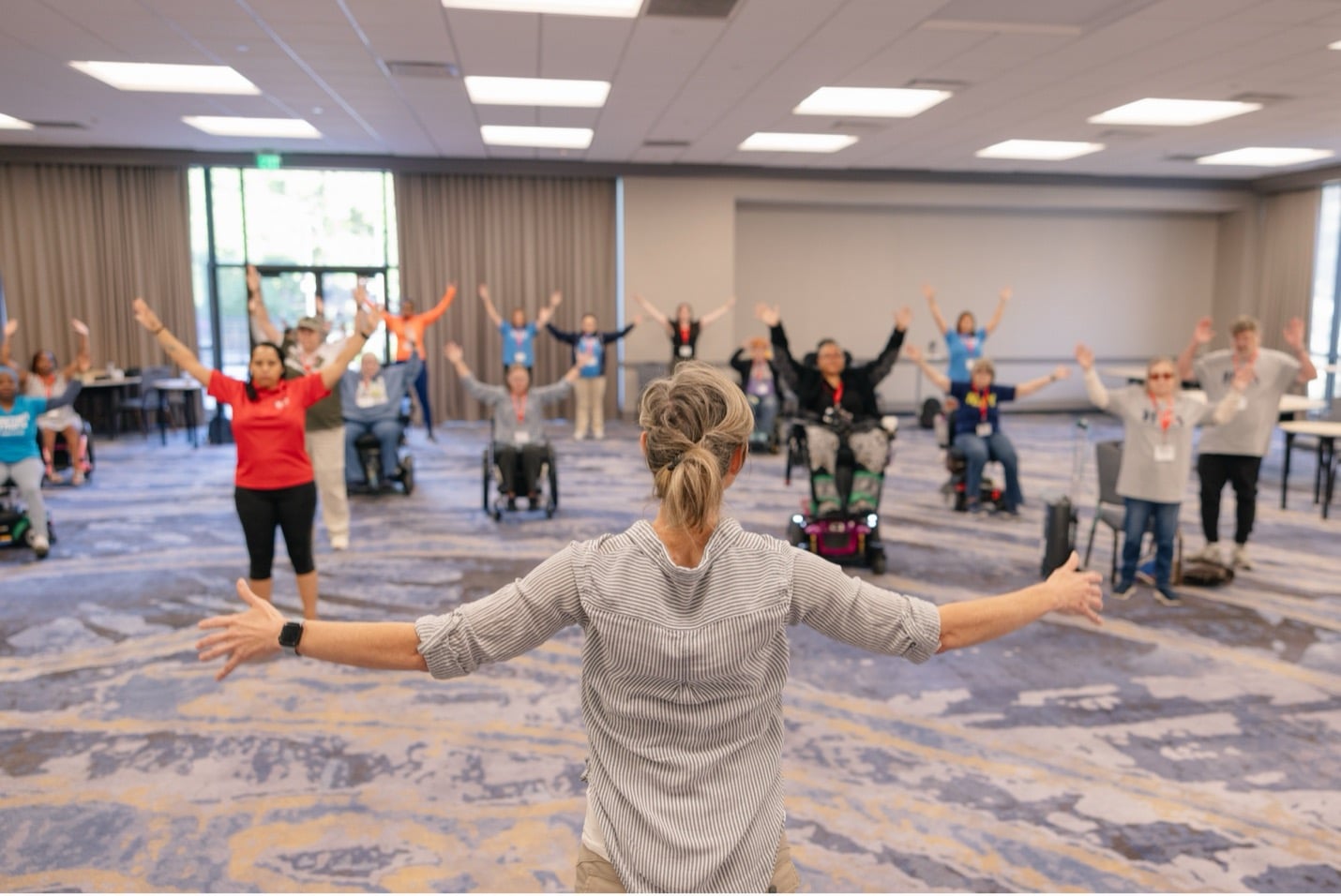 A group of people, some in wheelchairs, participate in an exercise or dance class in a large room, following an instructor at the front with arms outstretched.