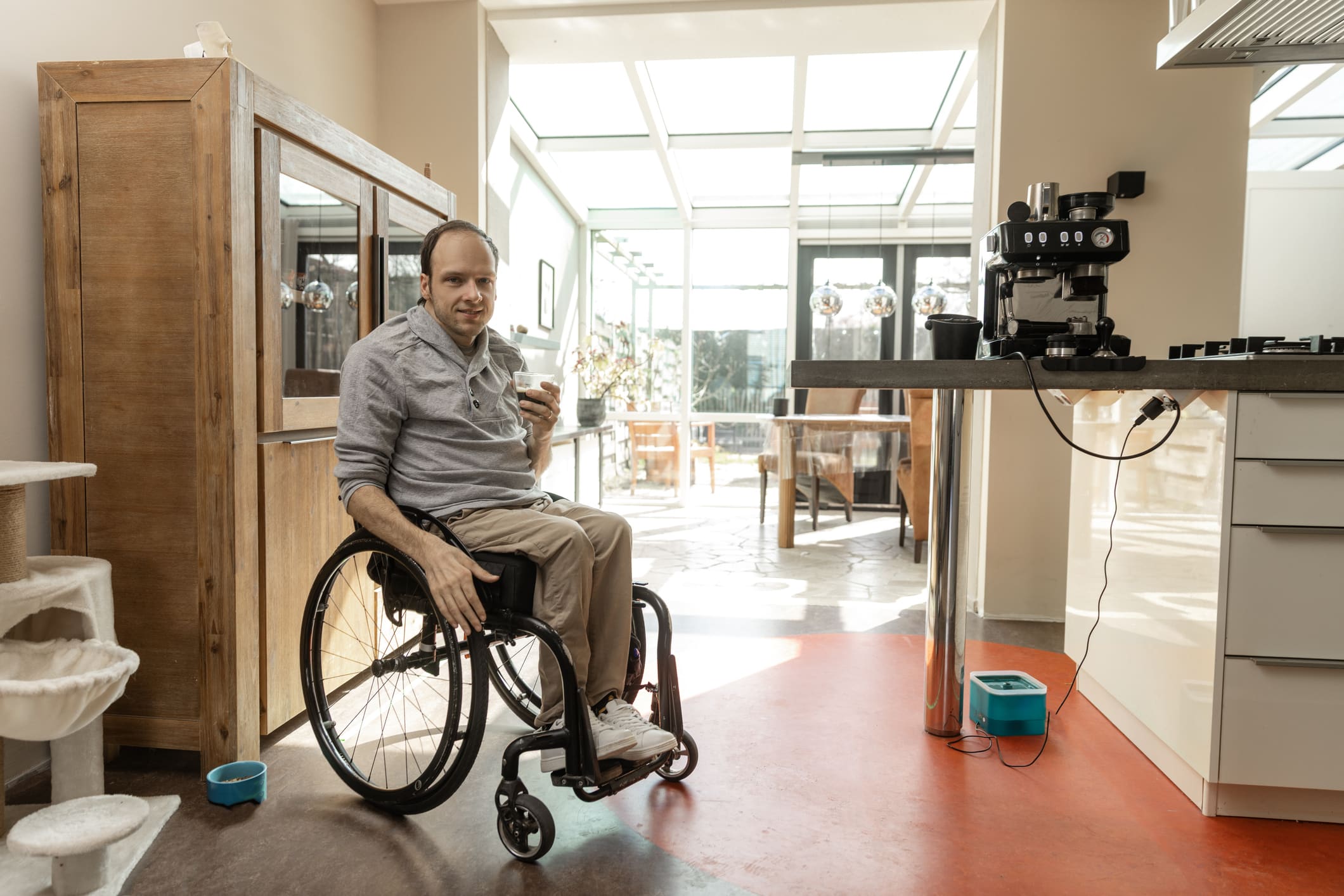 A man in a wheelchair holds a mug while sitting in a bright, modern kitchen with a coffee machine on the counter and sunlight streaming through large windows.