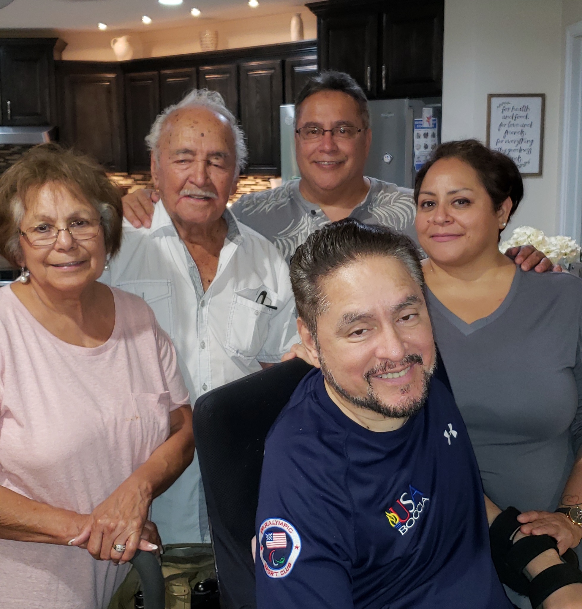 Five adults smile together in a cozy kitchen. One man is in a wheelchair at the front, and the others stand closely behind him, showing warmth and togetherness. The background features dark cabinets and soft lighting.