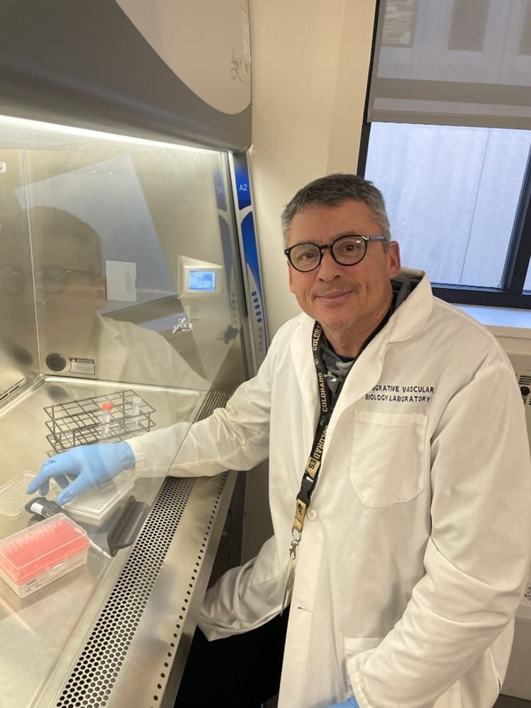 Dr. Christopher DeSouza, a scientist wearing glasses, a white lab coat, and blue gloves sits at a lab bench with laboratory equipment and red liquid-filled trays, smiling at the camera.