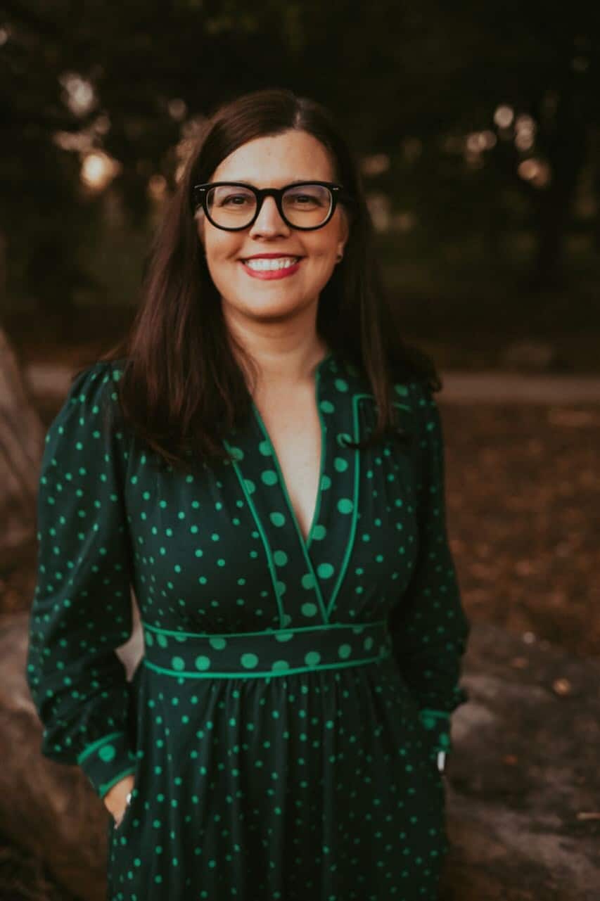 Dr. Laura Rice, a woman with long dark hair and glasses smiles outdoors, wearing a dark green dress with light green polka dots. The background is blurred with trees and a path.