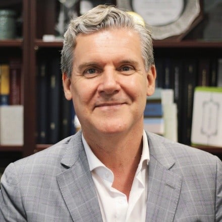 Dr. Michael Fehlings, a middle-aged man with gray hair, wearing a light gray checkered suit jacket and a white shirt, smiles at the camera. He is seated in front of a bookshelf filled with books and plaques.