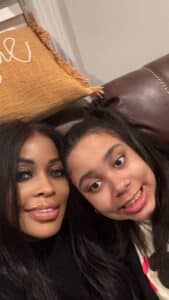 Two people with long dark hair sit close together, smiling for a selfie on a brown leather couch. Behind them is a tan pillow with white decorative script.