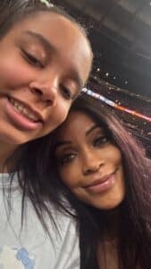 A woman and a young girl smile close together for a selfie inside an arena, with stadium lights and seating visible in the background.