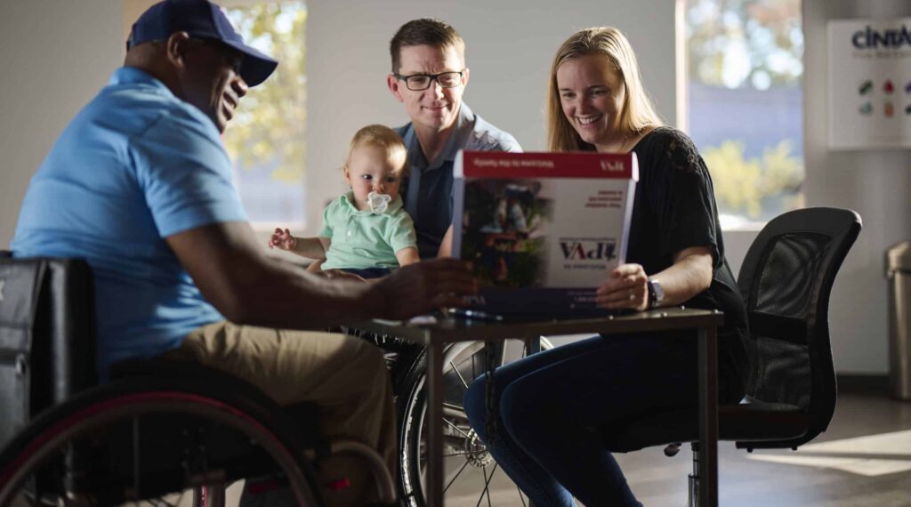A man in a wheelchair, a man with glasses holding a baby, and a smiling woman sit together at a table, looking at a brochure in a bright, modern room.
