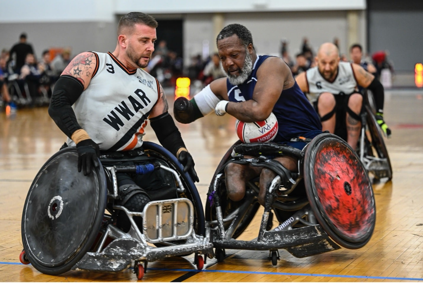 Two male wheelchair rugby players in action on a gym court, competing fiercely for the ball, with other players and spectators visible in the background.