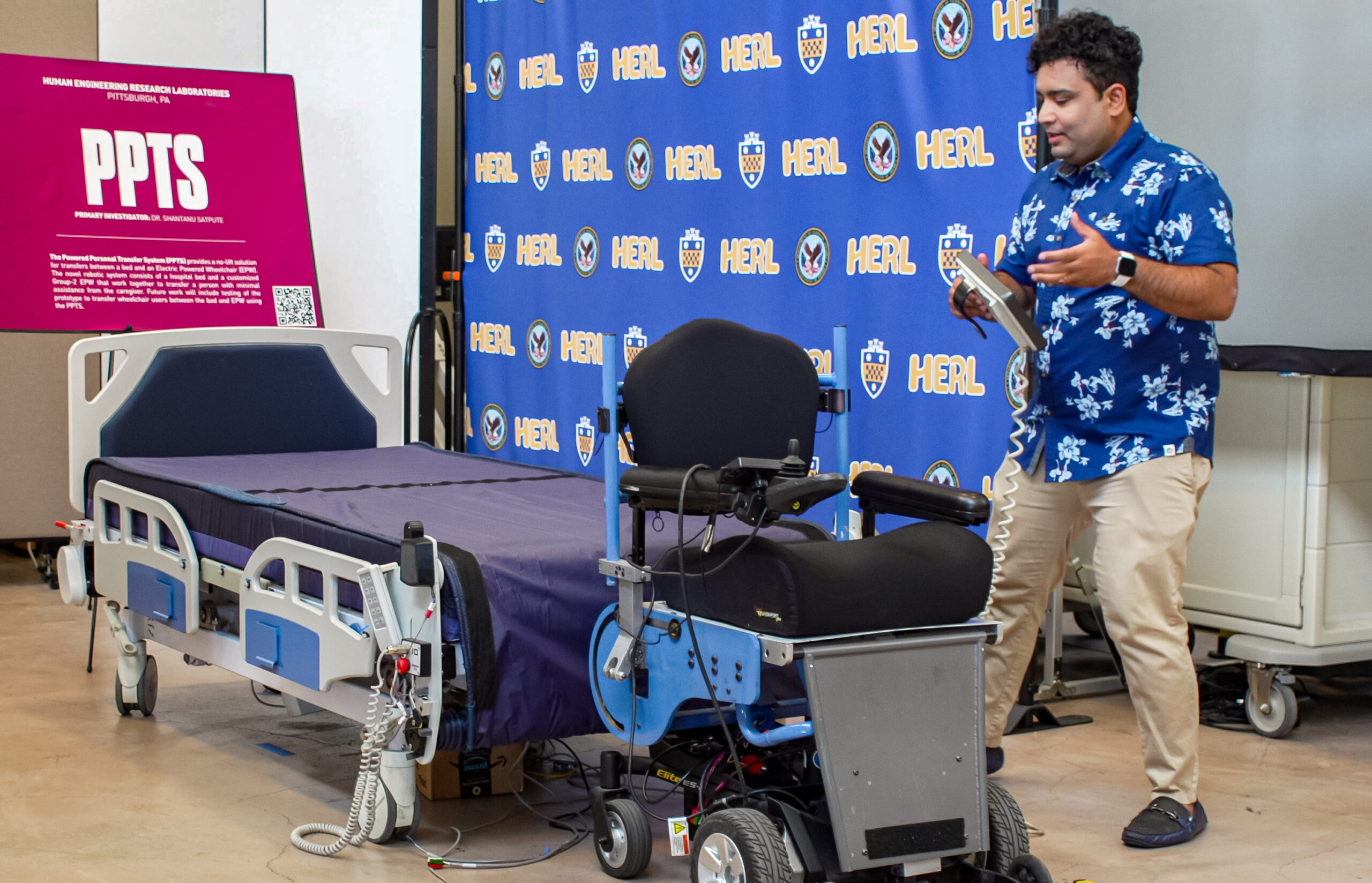 Dr Shantanu Satpute, a man in a blue button down shirt, stands next to a hospital bed and a large motorized wheelchair, gesturing as he presents. Behind him is a blue backdrop with "HERL" and logos, and a pink sign labeled "PPTS" is visible on the left.