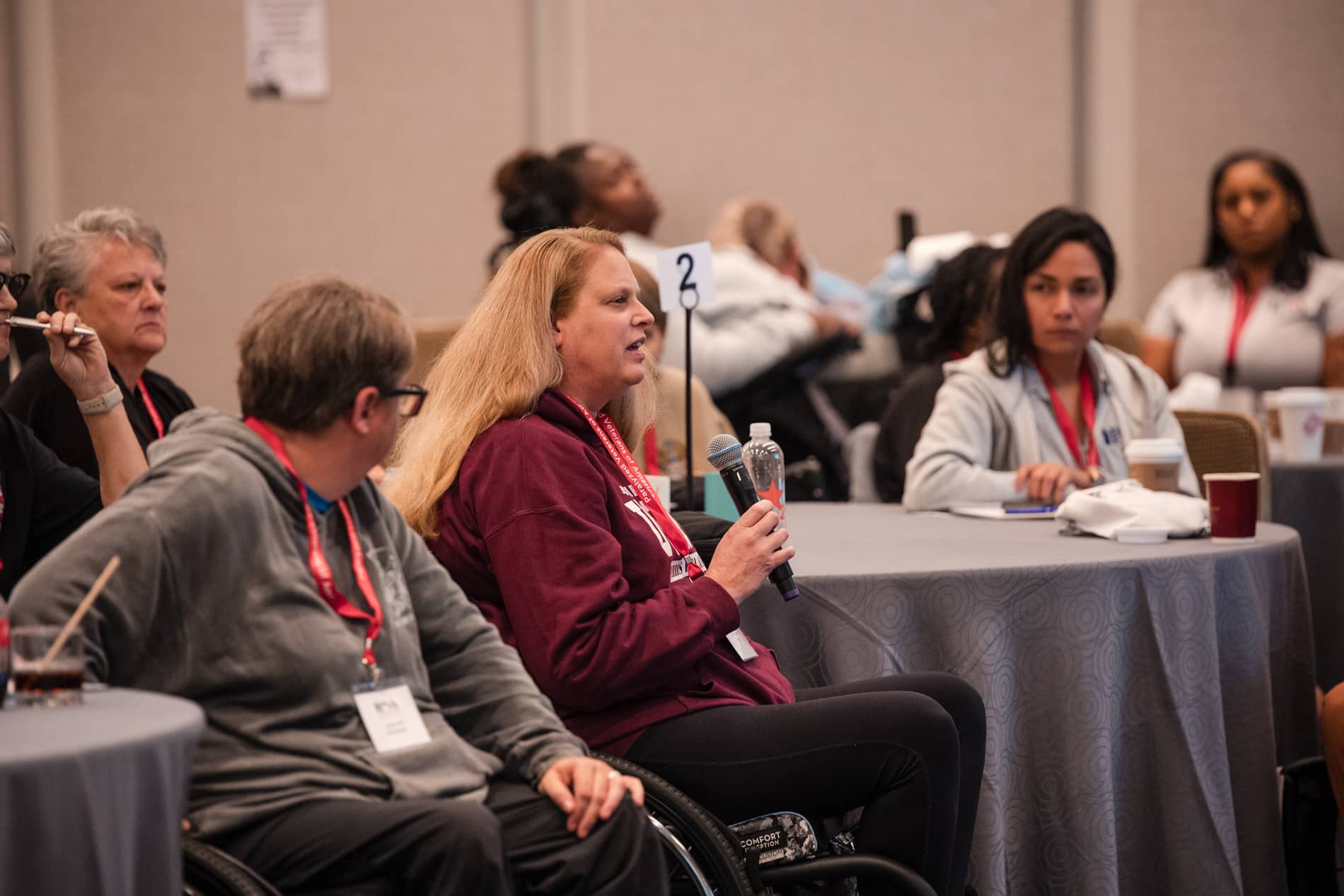 A woman in a maroon hoodie, seated in a wheelchair and holding a microphone, speaks at a conference. She is surrounded by others at round tables, with some people listening attentively.