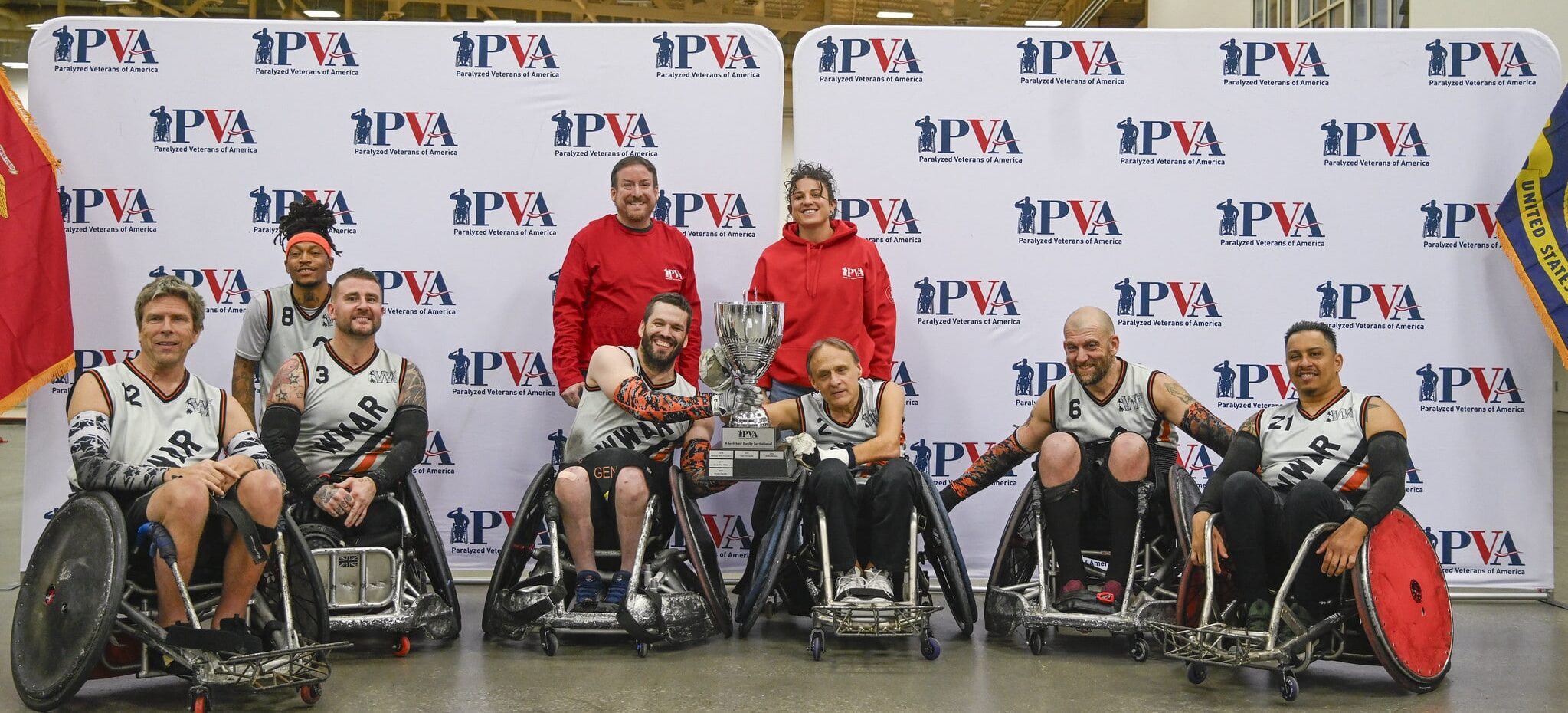 A wheelchair rugby team poses indoors with their trophy, flanked by coaches, in front of a backdrop featuring "PVA" logos. Most team members wear uniforms and smile at the camera.