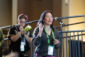A woman speaks into a microphone at a conference, wearing a name badge and green lanyard, while another person in the background takes notes in a notebook.