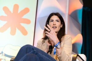 A woman with long dark hair speaks into a microphone on stage, gesturing with her hand. She wears a beige blazer, blue jeans, a watch, and blue wristbands, with colorful abstract shapes in the background.