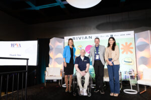 Four people pose and smile on stage at an event; one man is in a wheelchair. A Rivian banner and a slide reading "PVA Thank You" are visible in the background. Two women stand on either side of the two men.