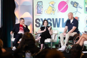 Three panelists sit onstage at SXSW, one woman speaking into a microphone between two men, one in a wheelchair. Behind them is a colorful illustrated backdrop with the SXSW logo and various cartoon icons. Audience is visible in foreground.