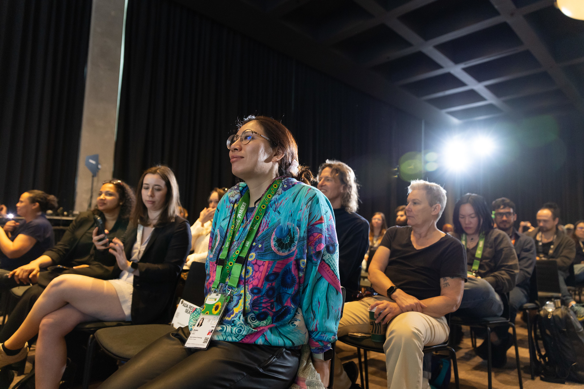 Four panelists sit on stage in front of a colorful illustrated backdrop at an event; one woman is speaking into a microphone, while the audience listens. One panelist uses a wheelchair.