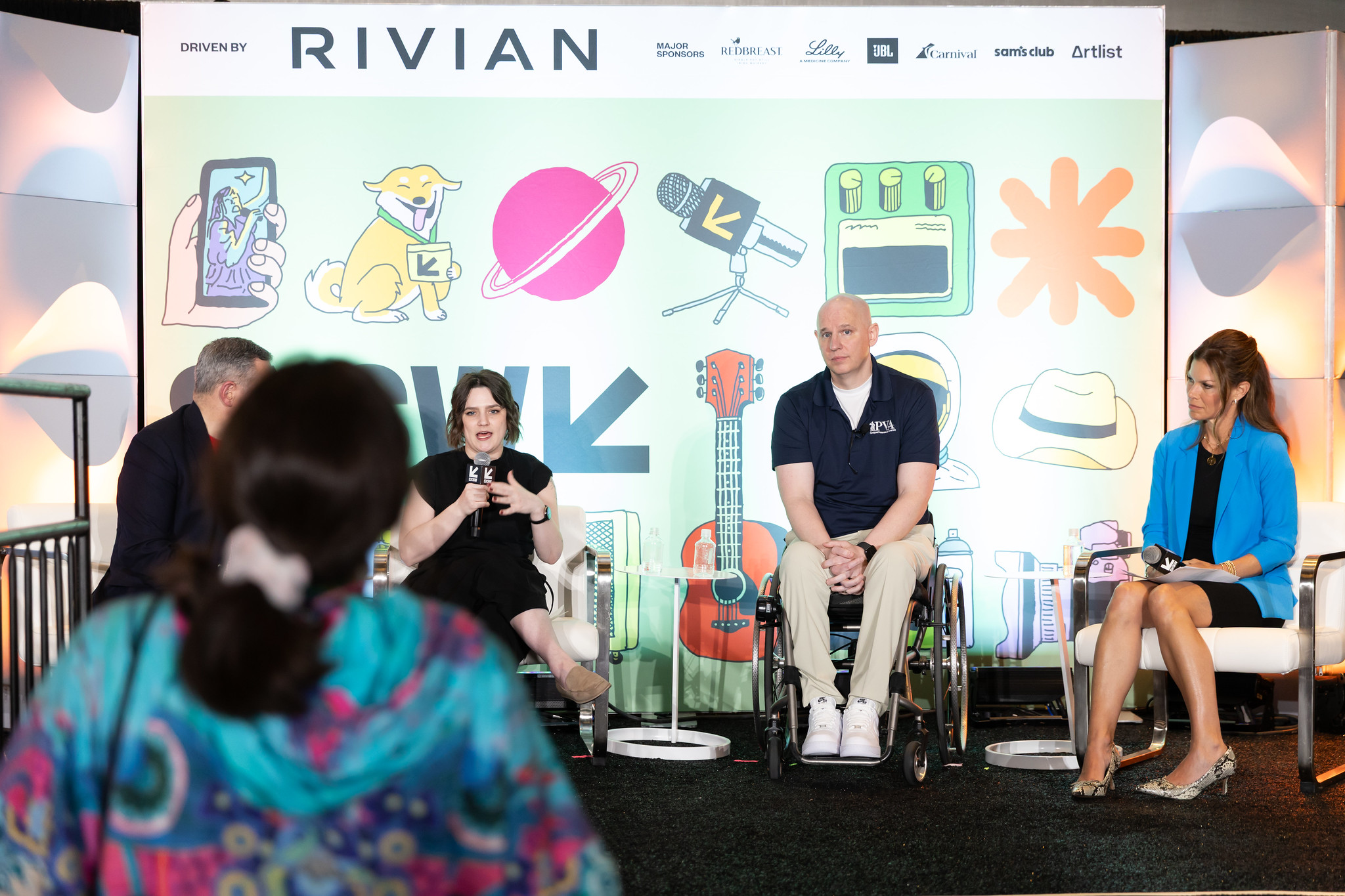 Four panelists sit on stage in front of a colorful illustrated backdrop at an event; one woman is speaking into a microphone, while the audience listens. One panelist uses a wheelchair.