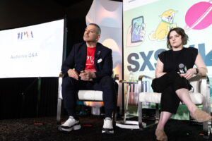 Two people sit on a stage during a panel discussion at SXSW. A screen beside them displays 