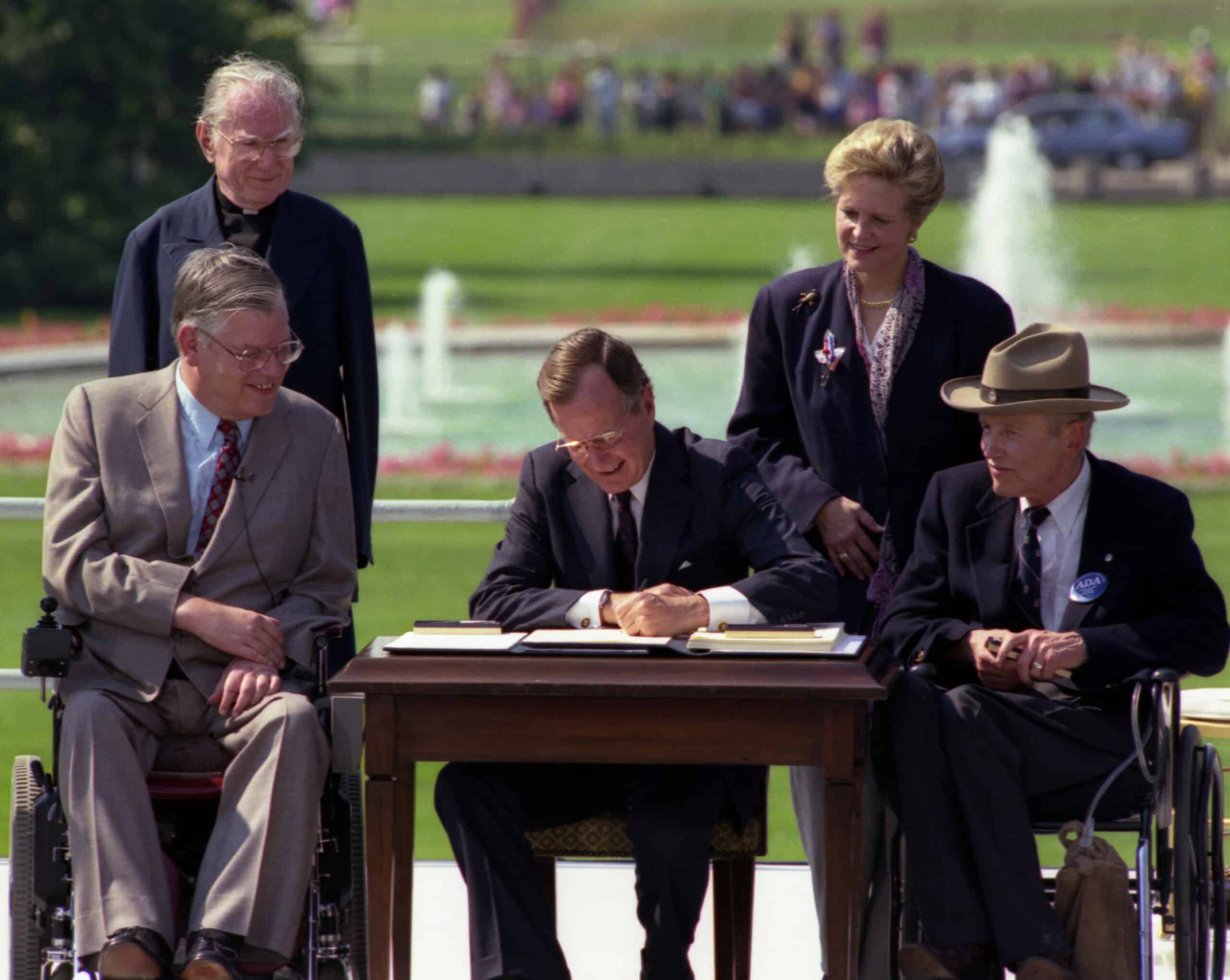 President George H.W. Bush, a man in a suit sits at a desk outdoors, signing a document, surrounded by four people, including two in wheelchairs. A fountain and a crowd are visible in the background.