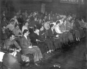 Black and white photo of a large group of men seated in rows on chairs, indoors, attending what appears to be a meeting or lecture. Many are wearing suits or uniforms, and some are looking forward attentively.