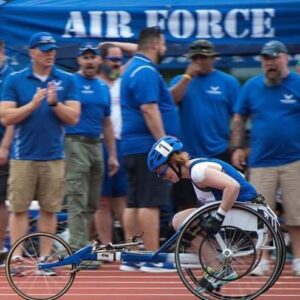 A wheelchair racer competes on a track in front of a group of people wearing blue shirts and a tent labeled 