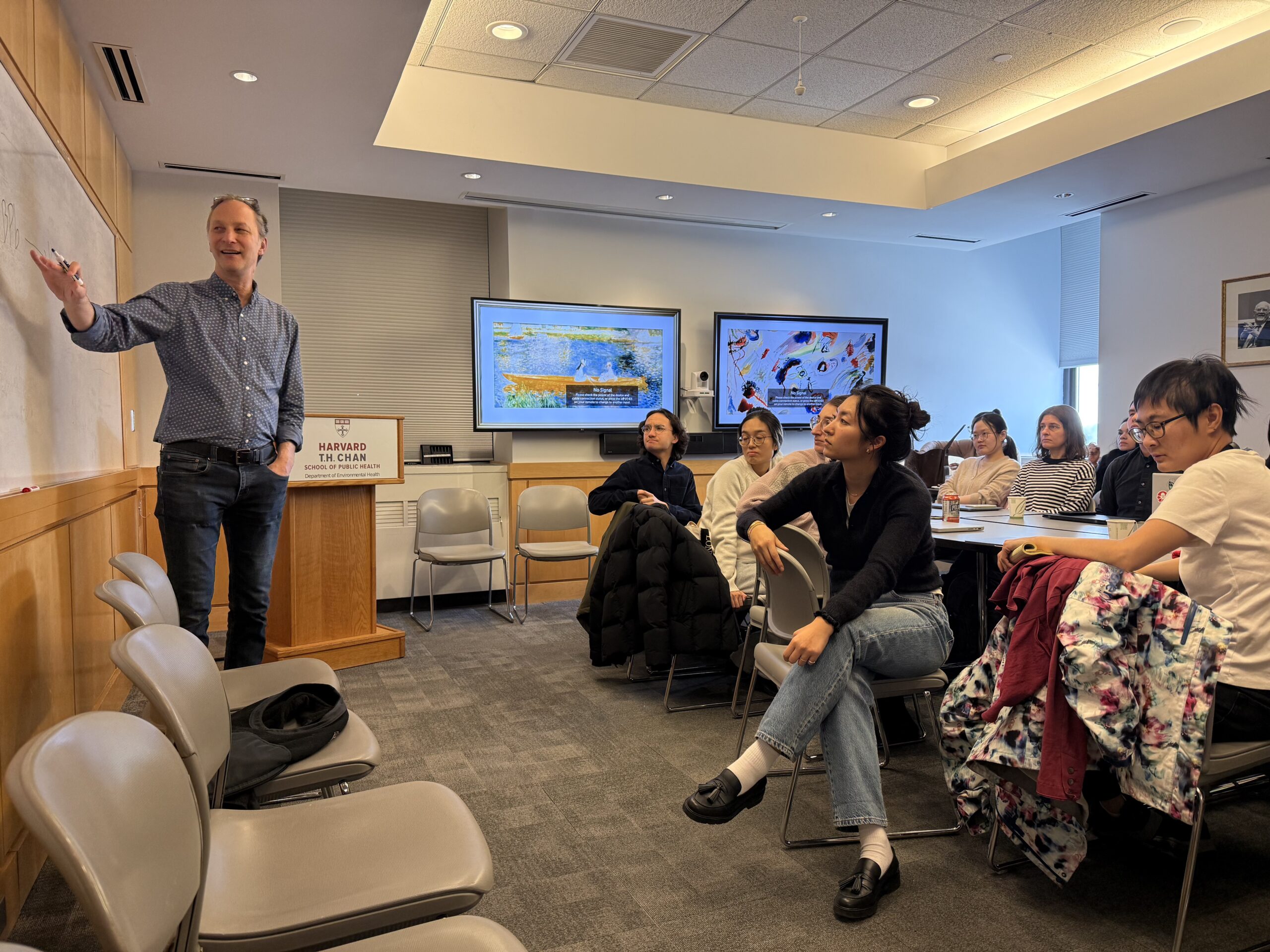 A man - Dr Marc Weisskopf - stands at a whiteboard giving a lecture to a group of attentive students seated in a classroom. Two large monitors display colorful visuals behind the students. The room is well-lit and modern.