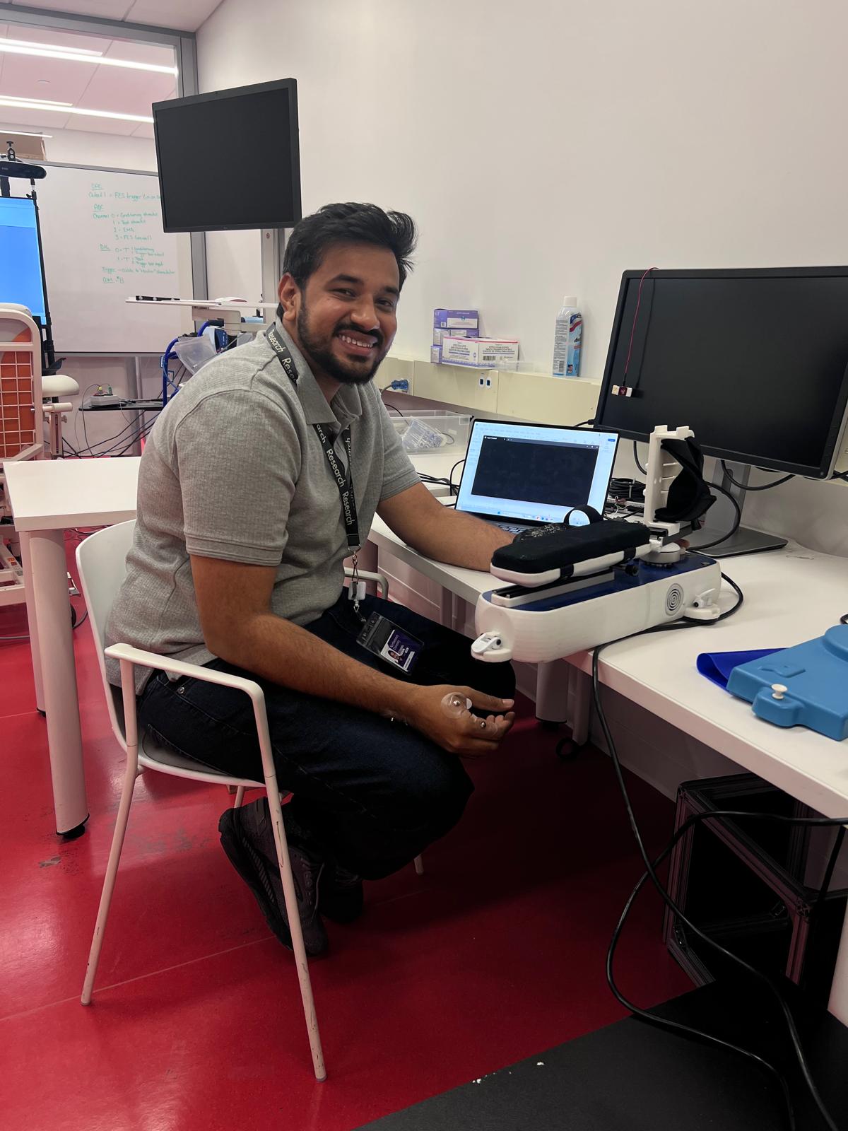 Dr. Aravind Nehrujee, a man in a grey polo, smiles while seated at a desk with electronic equipment, including a laptop and monitor, in a modern workspace with red floors and white walls.