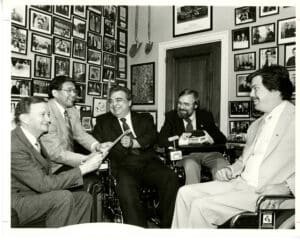 Five men in suits, some in wheelchairs, sit in a room filled with framed photos covering the walls. They are gathered in a friendly, lively conversation, sharing papers and smiling.