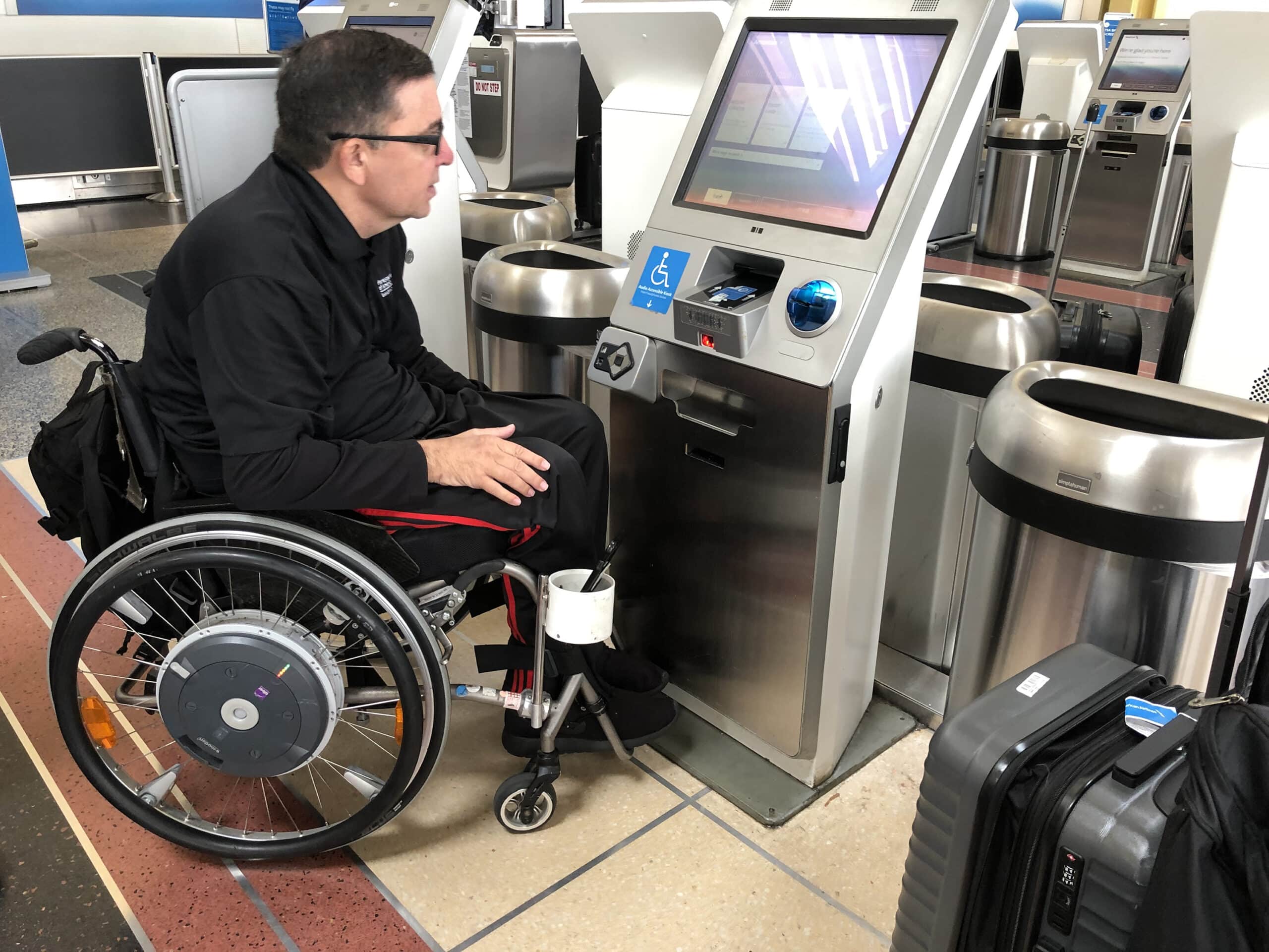 A man in a wheelchair uses an airport self-service kiosk, interacting with the touchscreen. A suitcase is nearby, and accessibility signage is visible on the kiosk.