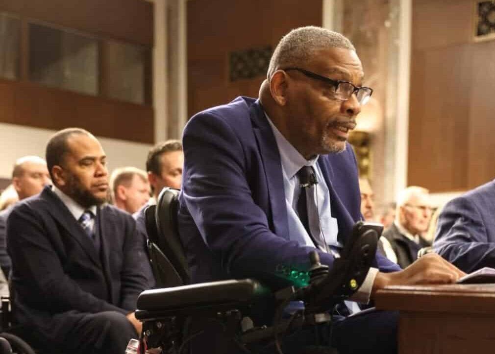 Robert Thomas, PVA National President, a man in a suit sits in a motorized wheelchair, attentively speaking at a formal hearing on Capitol Hill, in a wood-paneled room filled with other seated attendees.
