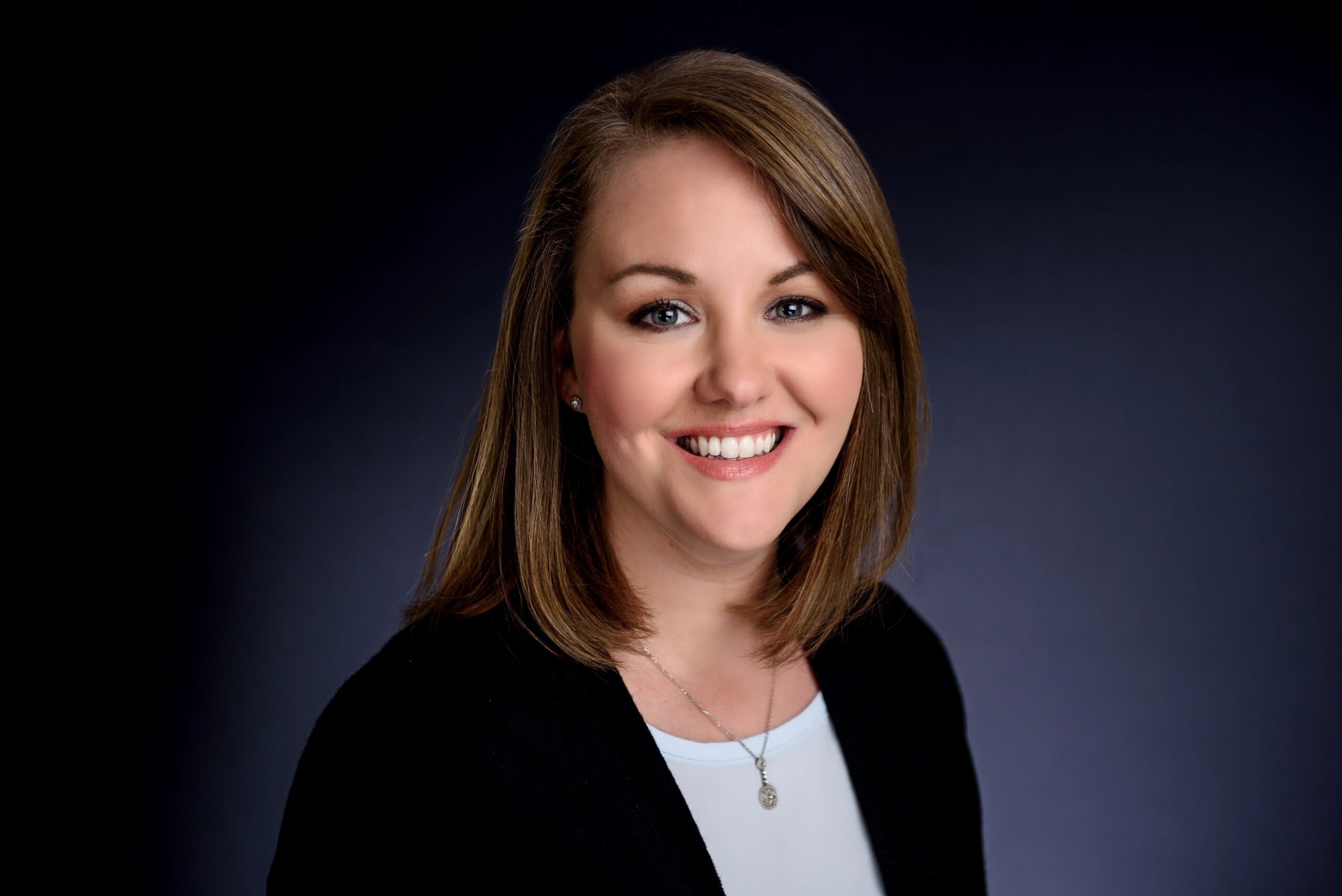 Dr. Brooks Wingo, a woman with straight, shoulder-length brown hair smiles at the camera. She is wearing a black blazer over a light blue top and a silver necklace, set against a dark blue background.