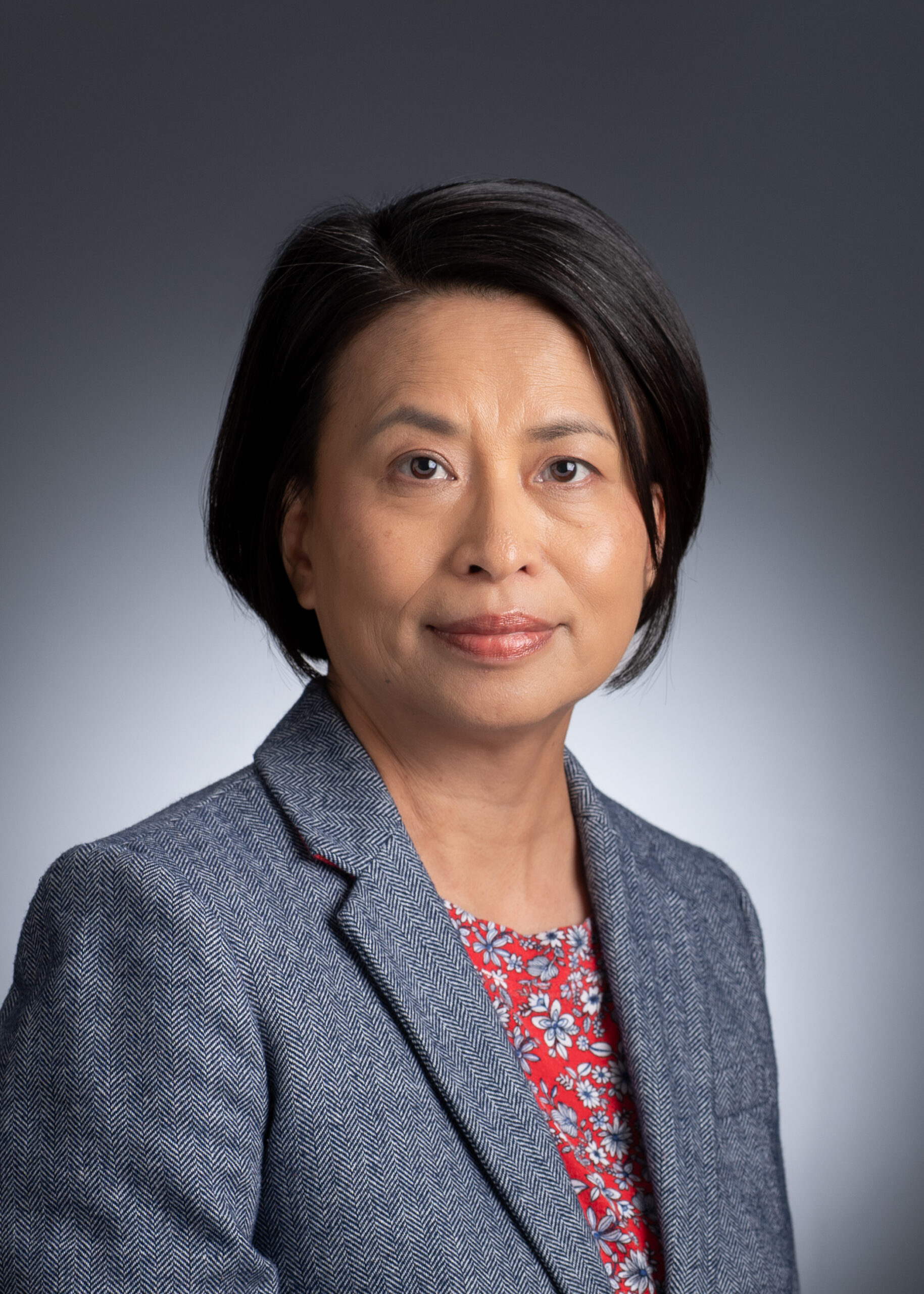 Dr. Yuying Chen, a woman with short black hair wearing a grey blazer over a red floral top poses for a professional portrait against a plain, gradient grey background.