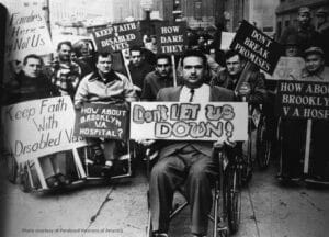 A group of men in wheelchairs protest on a city street, holding signs such as 