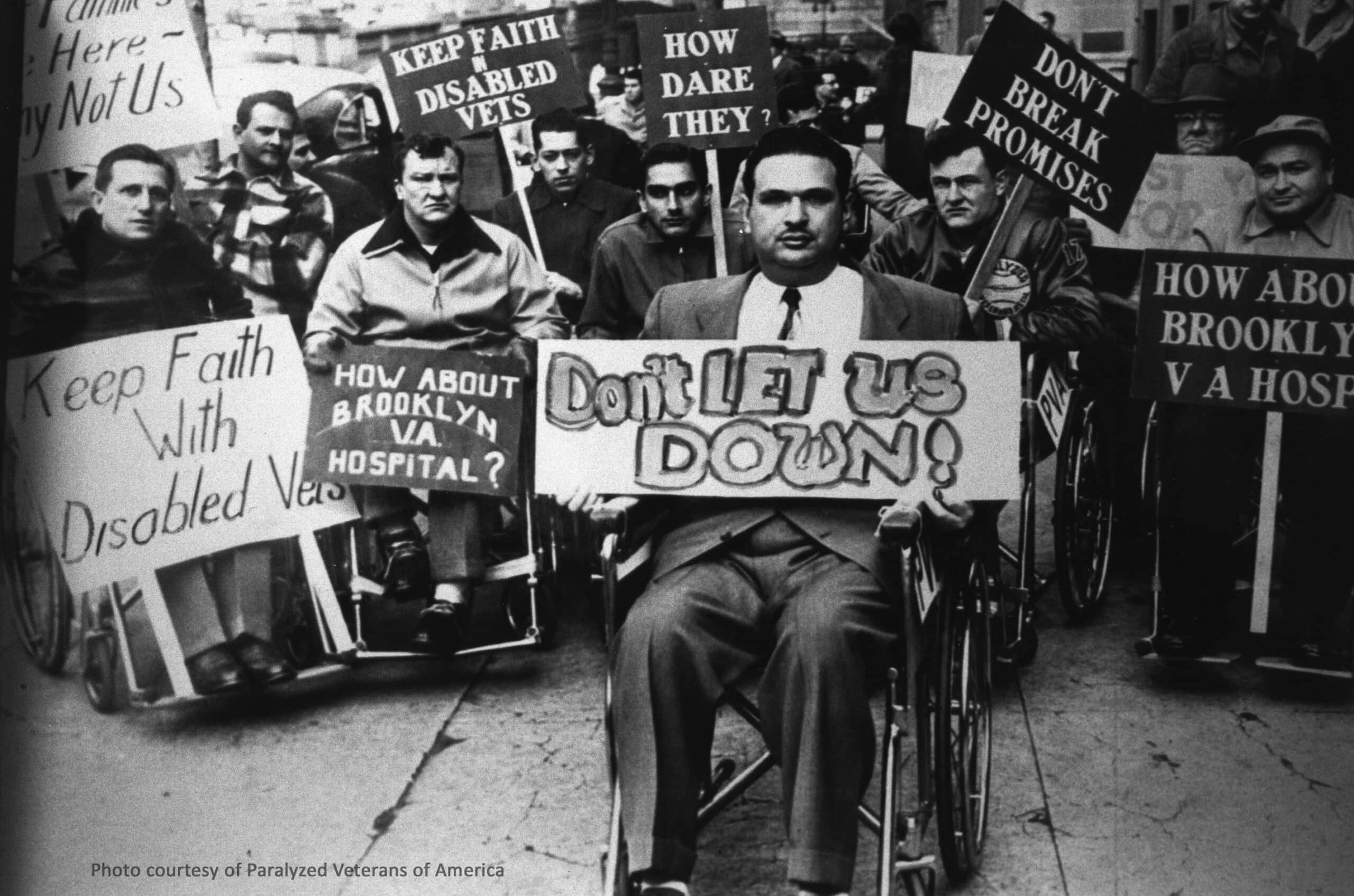 A group of men in wheelchairs protest on a city street, holding signs such as "Don't Let Us Down!", "Keep Faith With Disabled Vets," and "Don't Break Promises.